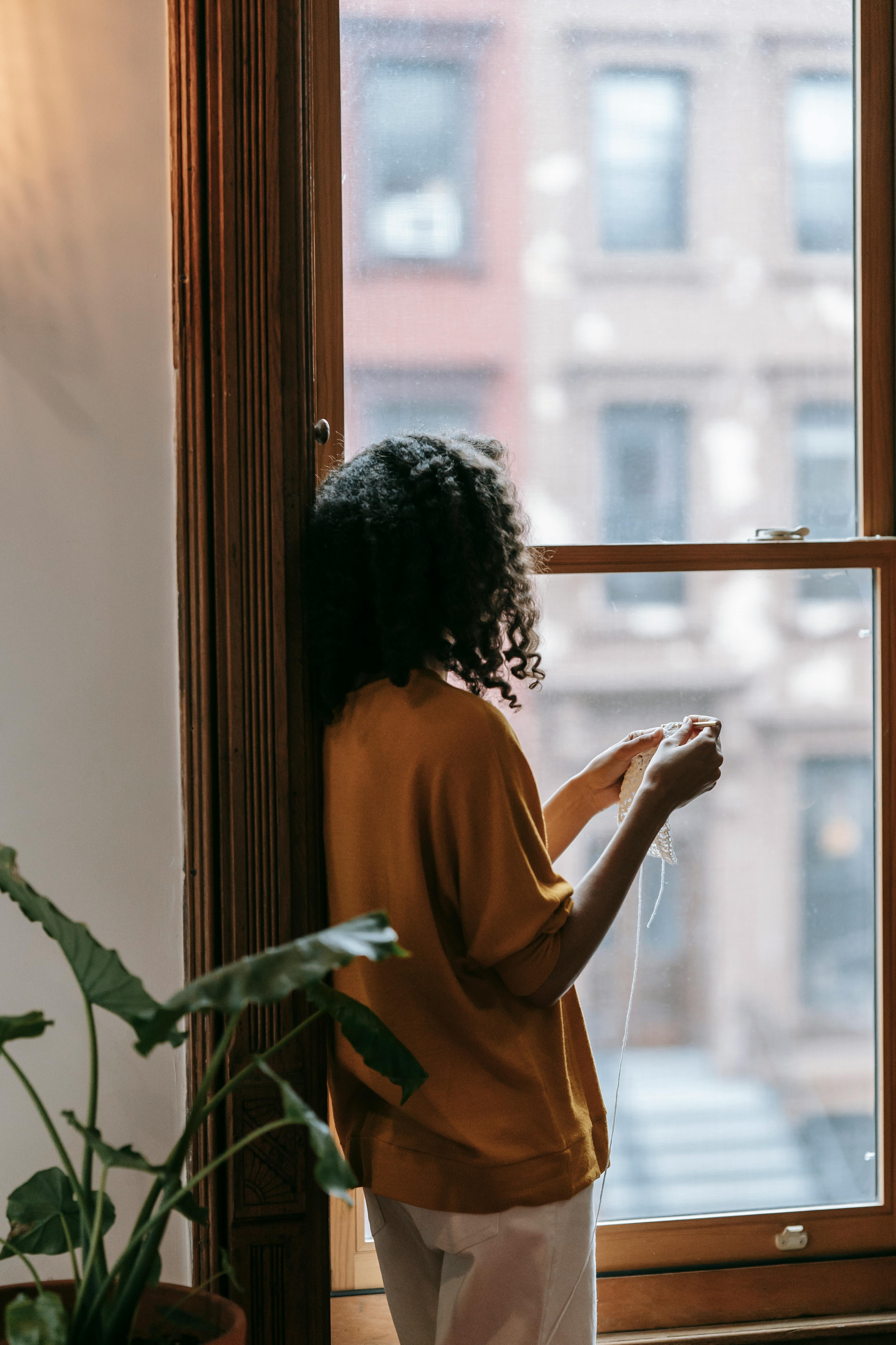 Black woman with natural hair standing at window holding curtain string looking outside in quiet contemplation with indoor plant nearby